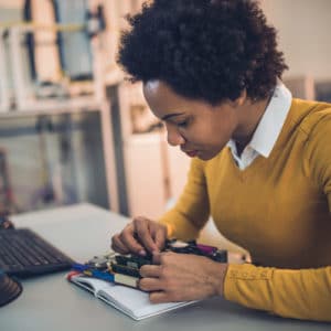 Female computer hardware engineer leans over desk to work on an electric circuit board project in an office