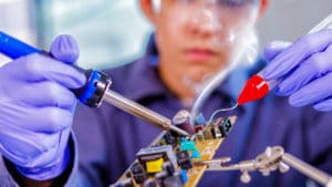 Female electronics engineer in a purple shirt and protective goggles and nitrile gloves solders component to circuit board