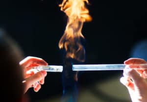 Woman holds two glass tubes with a flammable substance creating a flame in a dark room