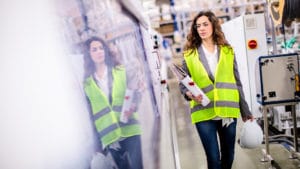 Female health & safety engineer in yellow vest holding a hardhat and radio inspecting an industrial work environment