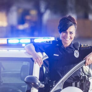 Woman police officer and detective smiles standing in her uniform next to her patrol car at night on a city street
