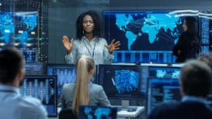 Woman information security analyst stands in front of a wall of digital global maps and speaks to fellow workers at their desks