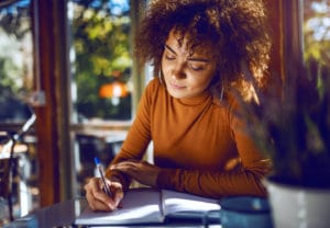 Young woman writing a story in a notebook while sitting near a window