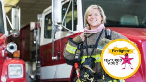 Female firefighter in protective safety coat and gloves stands in front of a red fire truck with white trim inside of a station